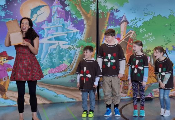 Fünf Kinder stehen auf einer Bühne, während eine Frau in einem roten Kleid mit einer Box posiert.