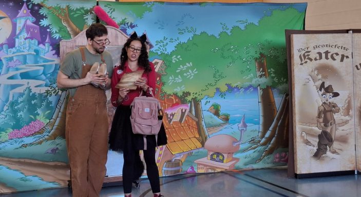 Eine verkleidete Person mit Rucksack und ein Mann stehen vor einem bunten Hintergrund.