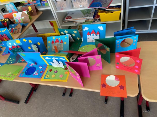 Bunte Bastelarbeiten mit Papier, Formen und Figuren auf Tischen in einem Klassenraum.