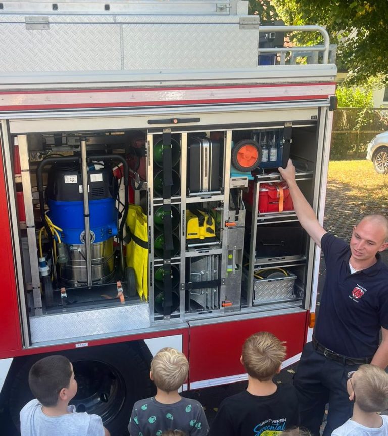 Feuerwehrmann erklärt Feuerwehrgeräte einer Gruppe neugieriger Kinder.